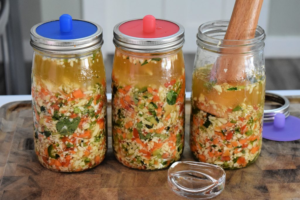 3 large mason jars full of colourful mixed vegetables in a brine solution. Two of the jars have silicone fermentation lids on them, and one is open with a wood tamper inside.