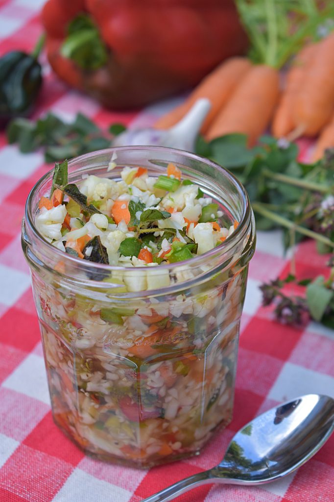 A jar of colorful fermented giardiniera sits on a red checkered cloth. Fresh vegetables are visible inside.