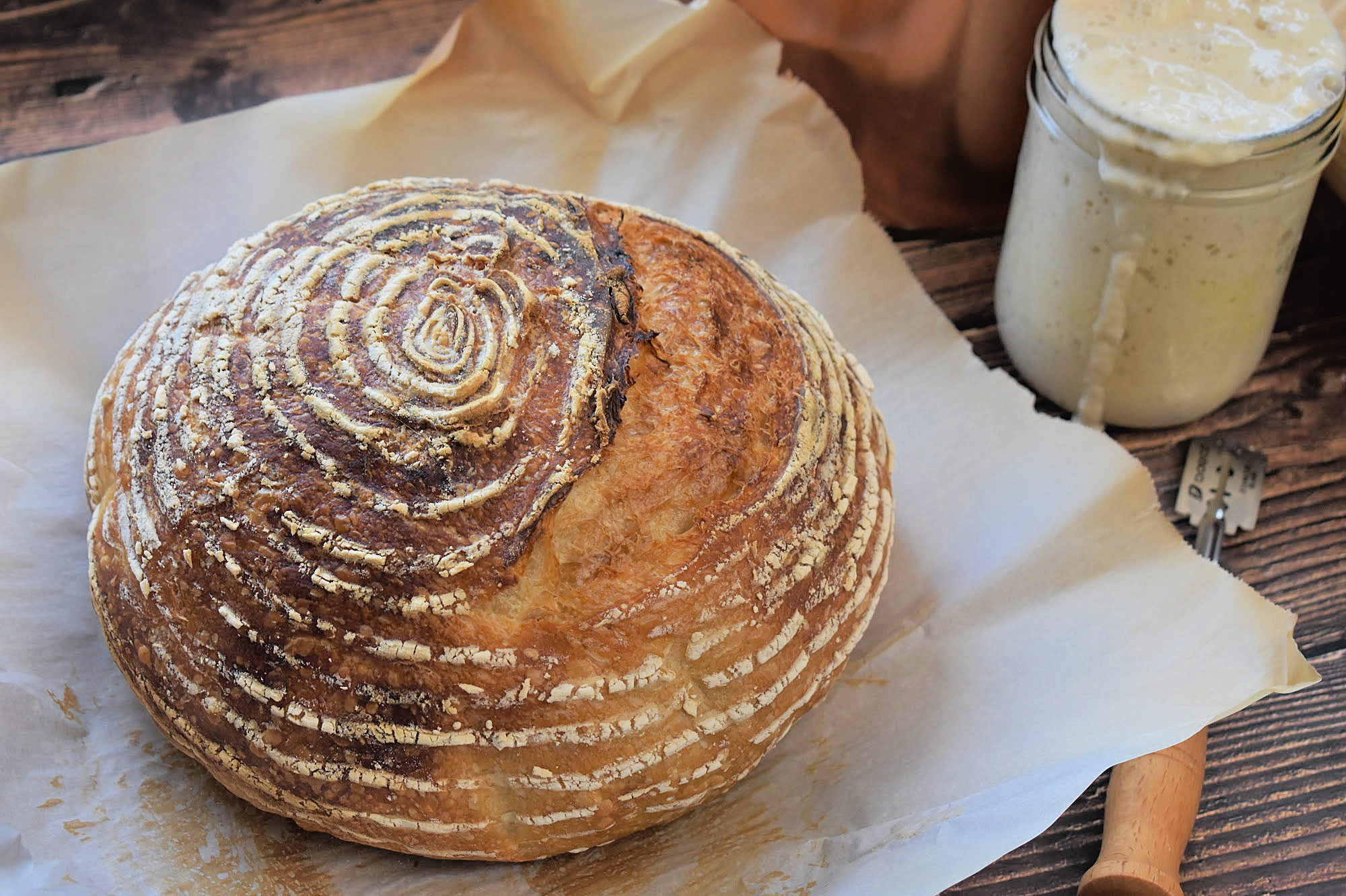 Easy Beginner Sourdough Bread Recipe Crusty artisan sourdough boule on parchment paper, next to a jar of active sourdough starter on a rustic wooden table.