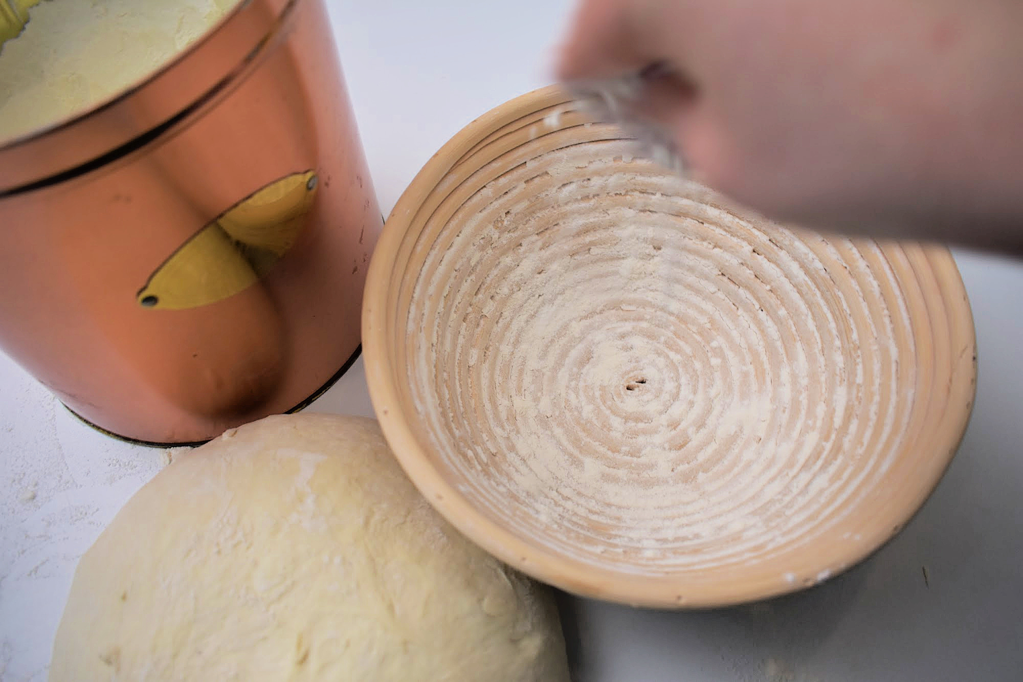 A hand flouring a round banneton basket beside a ball of sourdough and a copper flour canister.