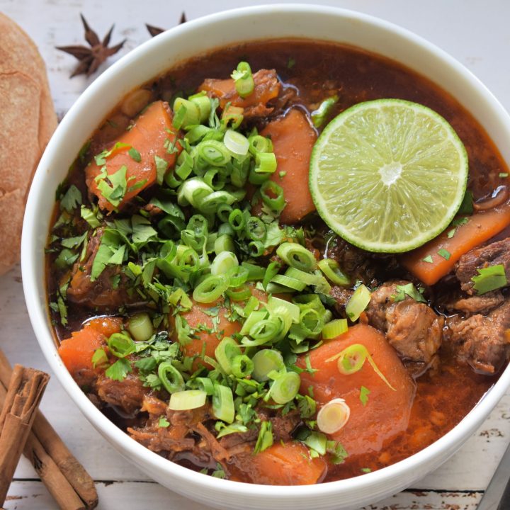 A bowl of hearty stew with chunks of meat and carrots, garnished with chopped green onions, cilantro, and a lime slice. Accompanied by a crusty roll, cinnamon sticks, and star anise on a wooden table. A spoon lies beside the bowl, creating a cozy, inviting ambiance.