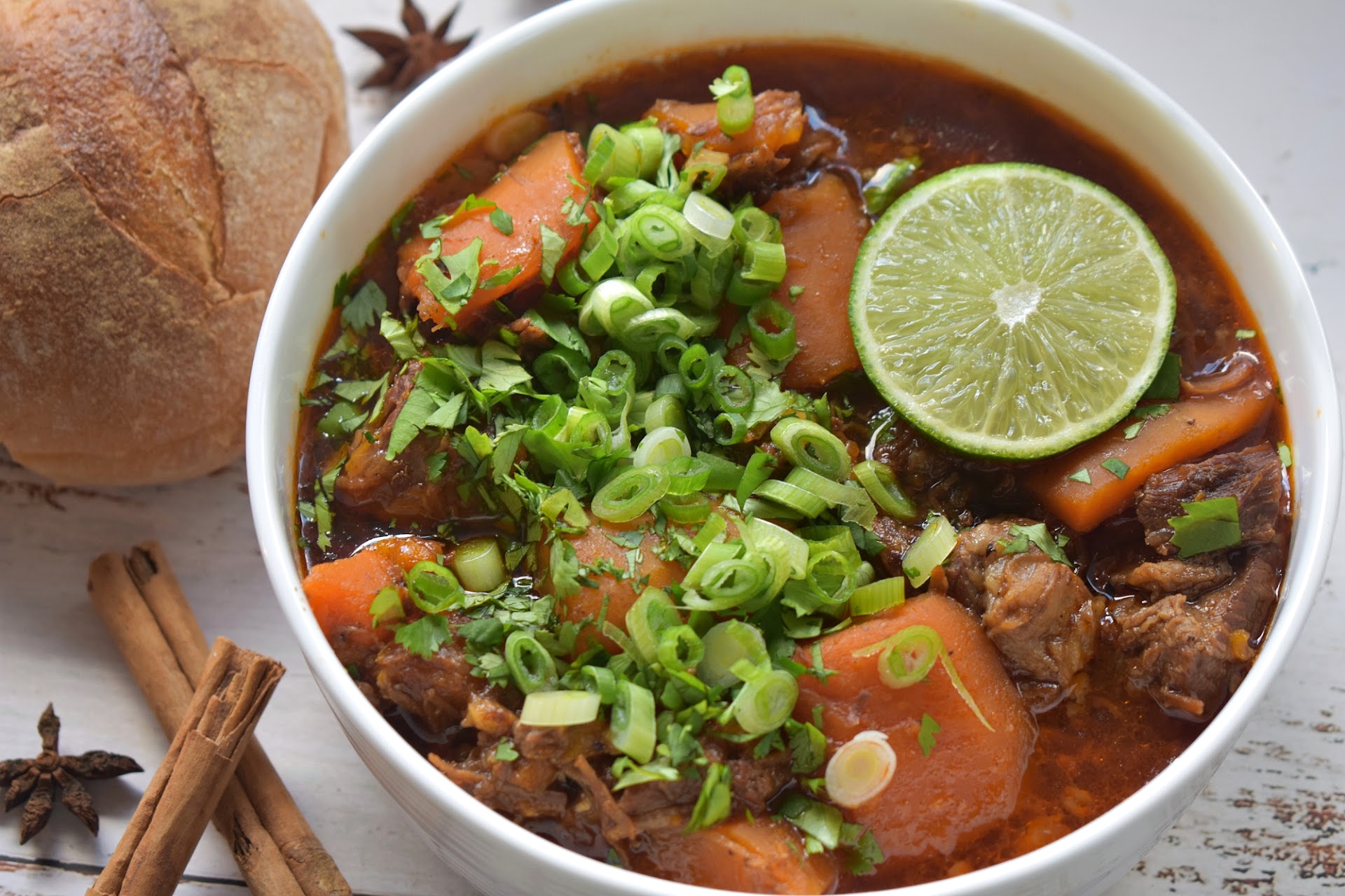 A bowl of vietnamese beef stew with chunks of meat and carrots, garnished with chopped green onions, cilantro, and a lime slice.