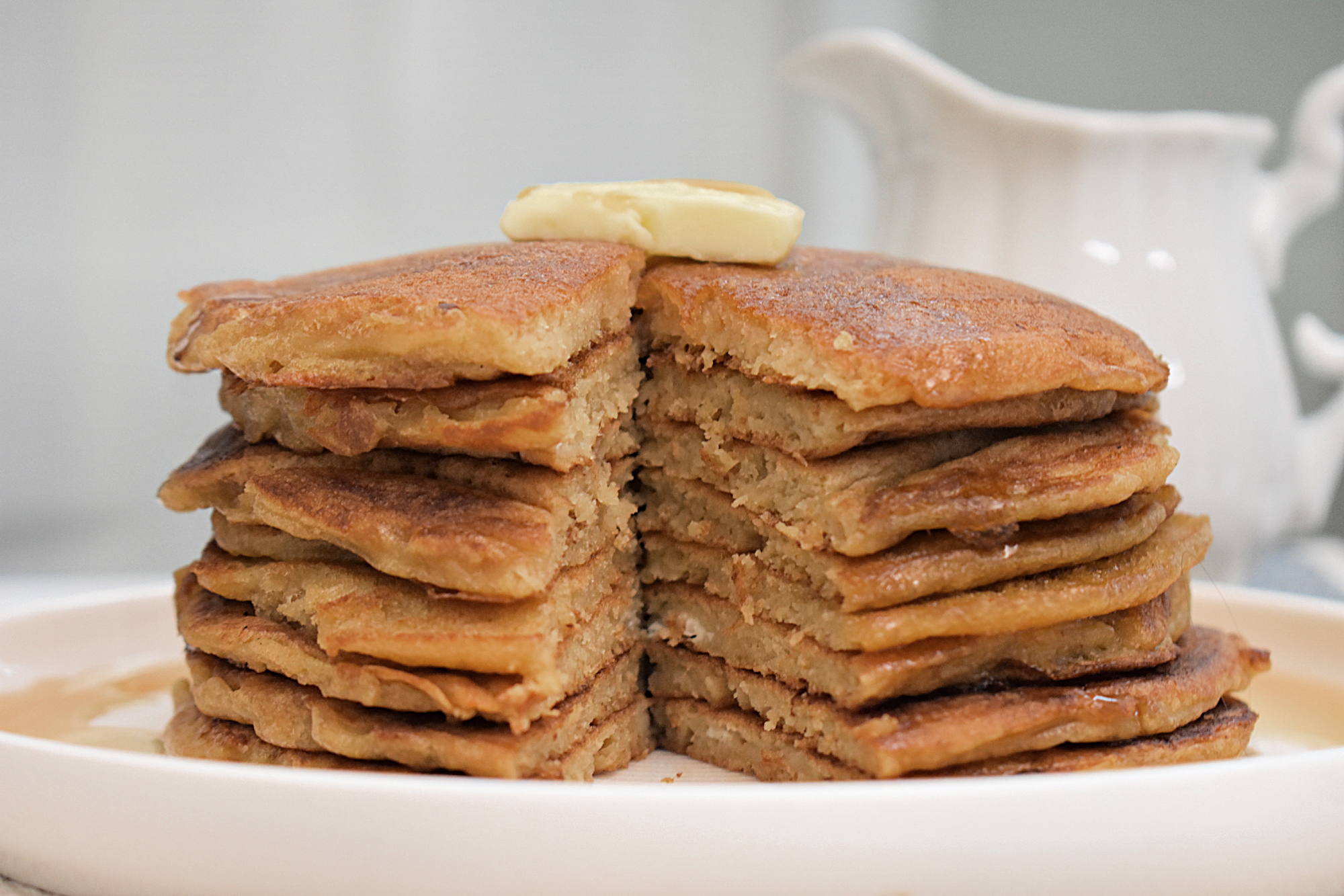 A fluffy stack of golden-brown sourdough discard pancakes with a wedge cut out. A pat of butter sits on top with syrup drizzled on top and on the surrounding plate.