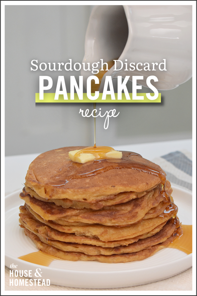 A stack of golden-brown sourdough discard pancakes on a white plate, topped with butter pats and syrup. A jug pours more syrup from above.