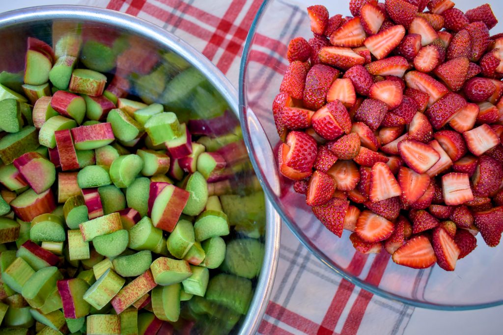 Two large bowls on a plaid tablecloth, one filled with chopped fresh rhubarb and the other full of sliced strawberries