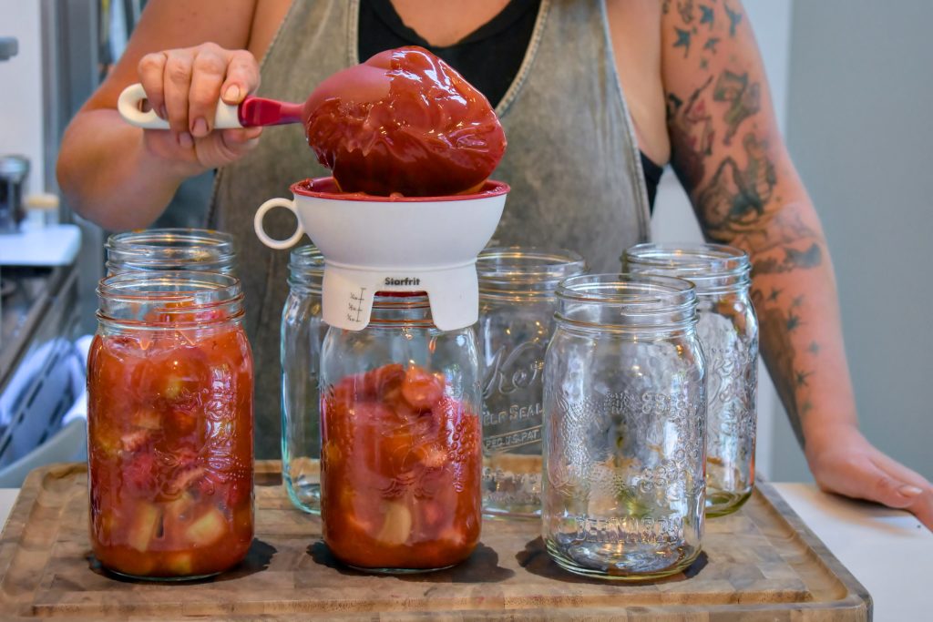A woman ladles prepared strawberry rhubarb pie filling into mason jars with a canning funnel