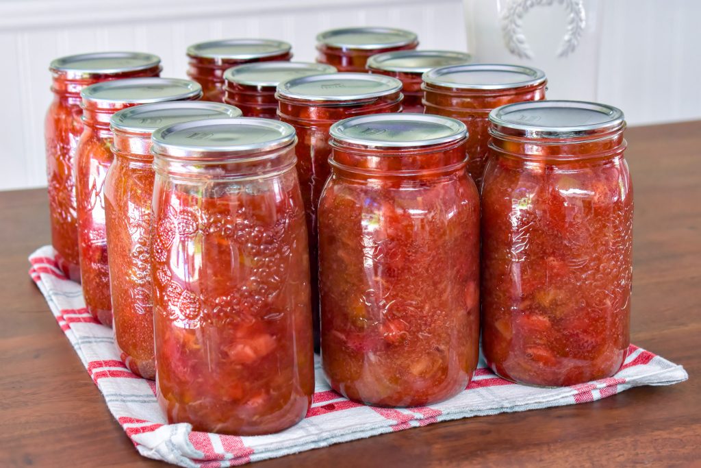 A dozen mason jars full of vibrant strawberry rhubarb pie filling sitting on a red checkered tea towel on top of a wooden tabletop