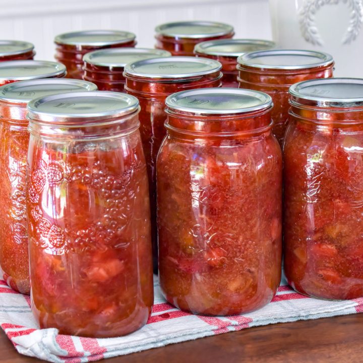 A dozen mason jars full of vibrant strawberry rhubarb pie filling sitting on a red checkered tea towel on top of a wooden tabletop