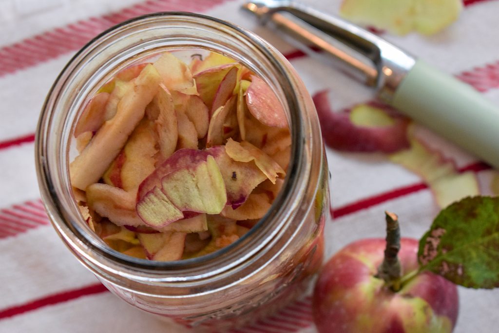 A top-down view of apple peels and skins in a glass mason jar sitting on a striped tea towel. A whole apple and peeler are visible in the background.
