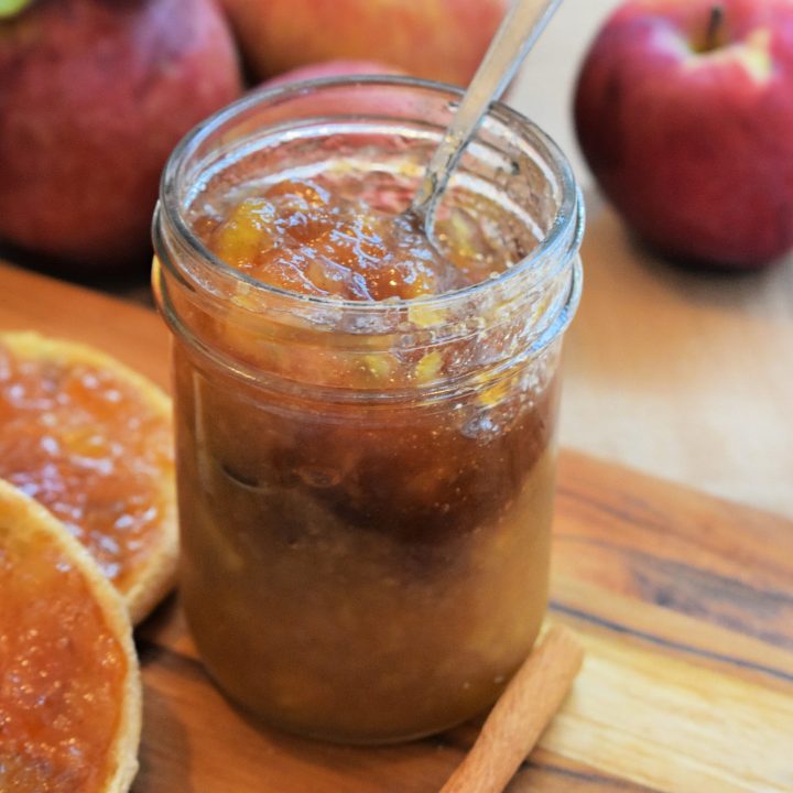 A jar of apple pie jam with the lid off. Chunks of apple are visible in the jam. Fresh apples are in the background and cinnamon sticks are in the foreground.